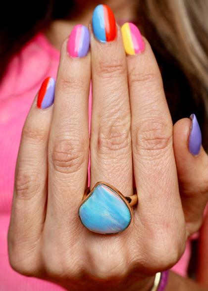 Close-up of a hand with colorful nail polish and a blue ring, against a blurred background.
